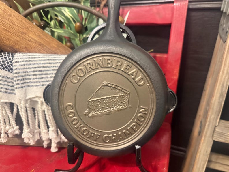 May include: A cast iron skillet with the words "CORNBREAD COOKOFF CHAMPION" embossed on the bottom. The skillet is black with a bronze-colored center, and a handle extends upwards. A slice of cornbread is depicted in the center.