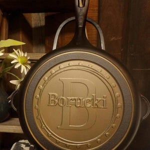 May include: A cast iron skillet with a handle and a decorative gold-colored center. The center features the name "Borucki" and a large letter "B". The skillet is displayed on a black metal stand.