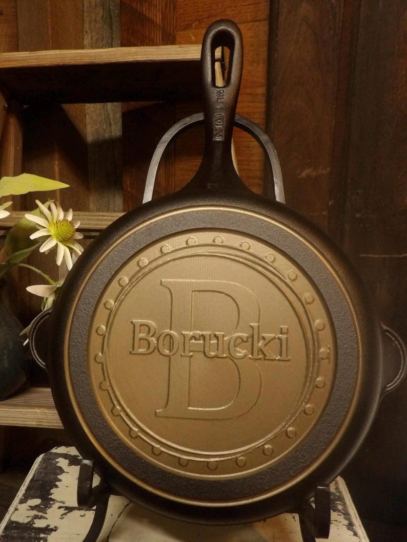 May include: A cast iron skillet with a handle and a decorative gold-colored center. The center features the name "Borucki" and a large letter "B". The skillet is displayed on a black metal stand.
