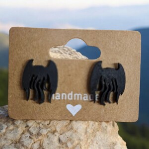 May include: A pair of black, 3D printed earrings shaped like a creature with wings. The earrings are displayed on a brown card with the word "handmade" printed on it.