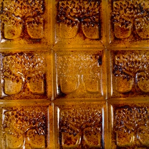 Pode incluir: Um conjunto de nove azulejos de vidro &acirc;mbar com um desenho em relevo de uma &aacute;rvore com dois troncos. Os azulejos est&atilde;o dispostos em uma grade de 3x3.