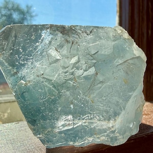 May include: A large, irregularly shaped, translucent aquamarine crystal. The crystal has a rough, textured surface with many facets, catching the light and creating a sparkling effect. The color is a pale, icy blue.