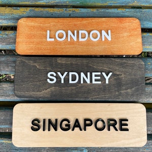 May include: Three wooden signs with the words "LONDON", "SYDNEY", and "SINGAPORE" written on them. The signs are stacked on top of each other, with the top sign being orange, the middle sign being dark brown, and the bottom sign being light brown.
