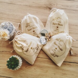 May include: Four beige drawstring bags with gold text and leaf designs. Each bag has a name, the words "Super Temain", and the date 08.05.2021. A small mirror and paper flower are next to the bags. A small succulent plant is in a white pot.