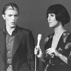 Può includere: Una fotografia in bianco e nero di due persone, un uomo e una donna, in piedi uno accanto all'altra. L'uomo indossa una giacca di un completo e una camicia abbottonata. La donna indossa un abito nero a maniche lunghe con un motivo floreale. Tiene un microfono.