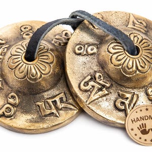 May include: Two antique brass tingsha cymbals with a dark leather strap. Each cymbal features embossed floral and geometric designs, and is stamped with symbols. A small, round "HANDMADE" sticker is attached.