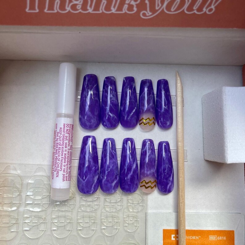 Op de afbeelding: Een set opzetnagels in een doos. De nagels zijn gemarmerd paars met witte adering. E&eacute;n nagel heeft een gouden zigzagontwerp. Ook inbegrepen zijn nagellijm, nagelvoorbereidingspads en een houten cuticle stick.