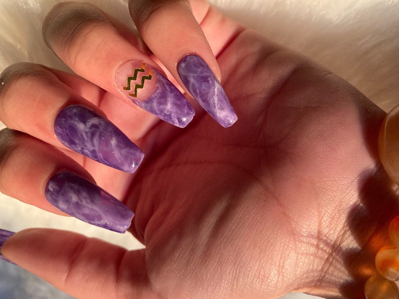 Op de afbeelding: Close-up van een hand met lange, vierkante, paarse acryl nagels met marmer effect. E&eacute;n nagel heeft een gouden zigzag ontwerp. De nagels hebben een glanzende afwerking en de hand is versierd met een armband.