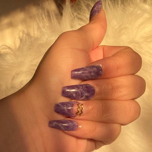 Op de afbeelding: Een hand met lange, coffin-vormige, paarse acryl nagels met marmer effect. E&eacute;n nagel heeft een goudkleurig zigzag ontwerp. De nagels zijn goed verzorgd en glanzend. De achtergrond is een zachte, witte, pluizige textuur.