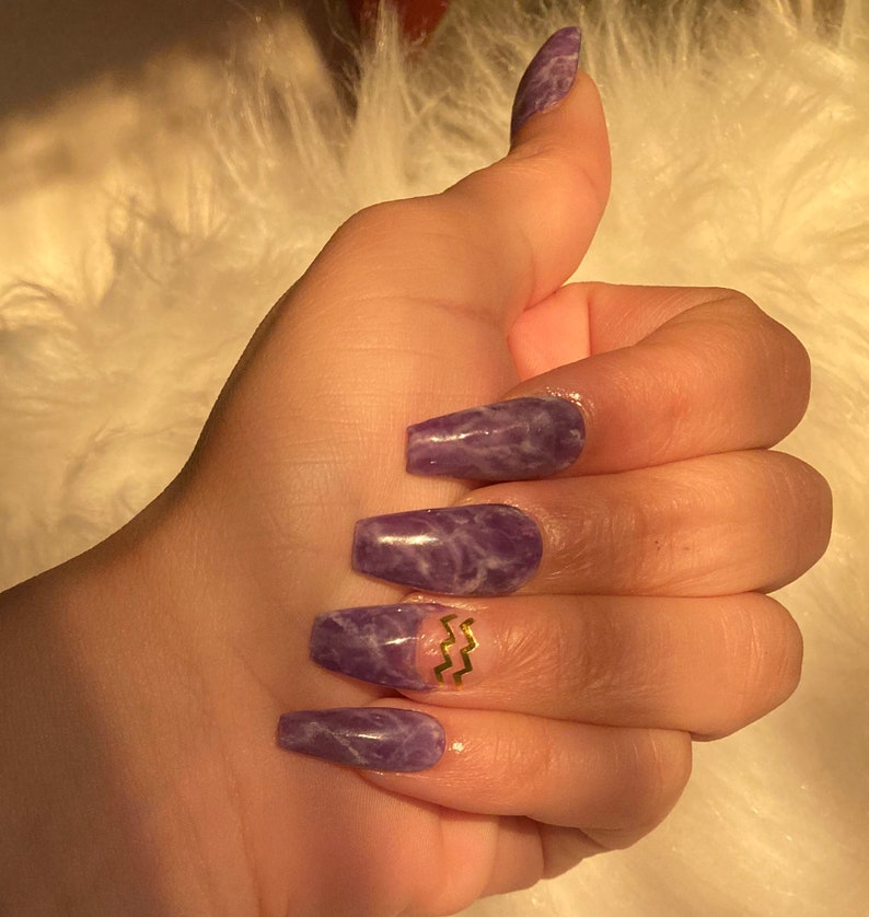 Op de afbeelding: Een hand met lange, coffin-vormige, paarse acryl nagels met marmer effect. E&eacute;n nagel heeft een goudkleurig zigzag ontwerp. De nagels zijn goed verzorgd en glanzend. De achtergrond is een zachte, witte, pluizige textuur.