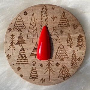 Puede incluir: Una sola uña roja vibrante sobre un posavasos de madera. El posavasos está decorado con árboles de Navidad y copos de nieve grabados. La uña es larga y puntiaguda, con un acabado brillante. El tema general es festivo y navideño.