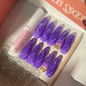 Op de afbeelding: Een set lange, rechthoekige persnagels met een paars marmerpatroon en gouden zigzagaccenten. De set bevat nagellijm en een houten cuticle pusher. De nagels zijn ongeveer 2 cm breed.