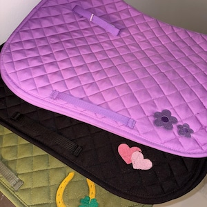 May include: A stack of quilted horse saddle pads in purple, black, and olive green. The purple pad has flower appliqués and a strap. The black pad has heart appliqués. The olive green pad has a horseshoe and clover appliqué.