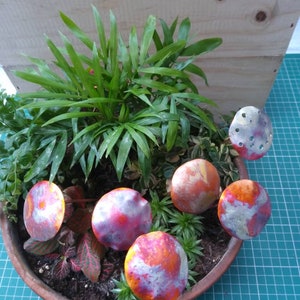 May include: Five decorative metal mushrooms with a red, orange and white paint design. The mushrooms are arranged in a potted plant with green leaves.