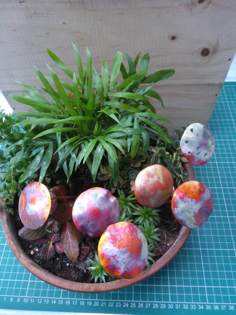 May include: Five decorative metal mushrooms with a red, orange and white paint design. The mushrooms are arranged in a potted plant with green leaves.