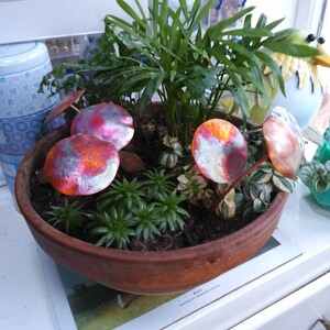 May include: A terracotta pot containing a variety of green plants and small copper mushrooms with orange and red accents.