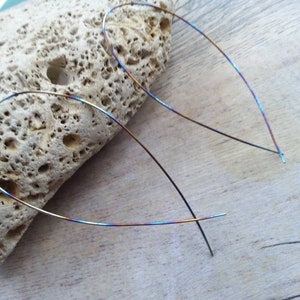 May include: A pair of thin, iridescent hoop earrings made of metal. The earrings are a rainbow of colours, including blue, green, gold, and purple.