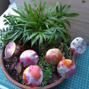 May include: Five small, metallic, mushroom-shaped ornaments in shades of orange, pink, and white, arranged in a terracotta pot with green plants.