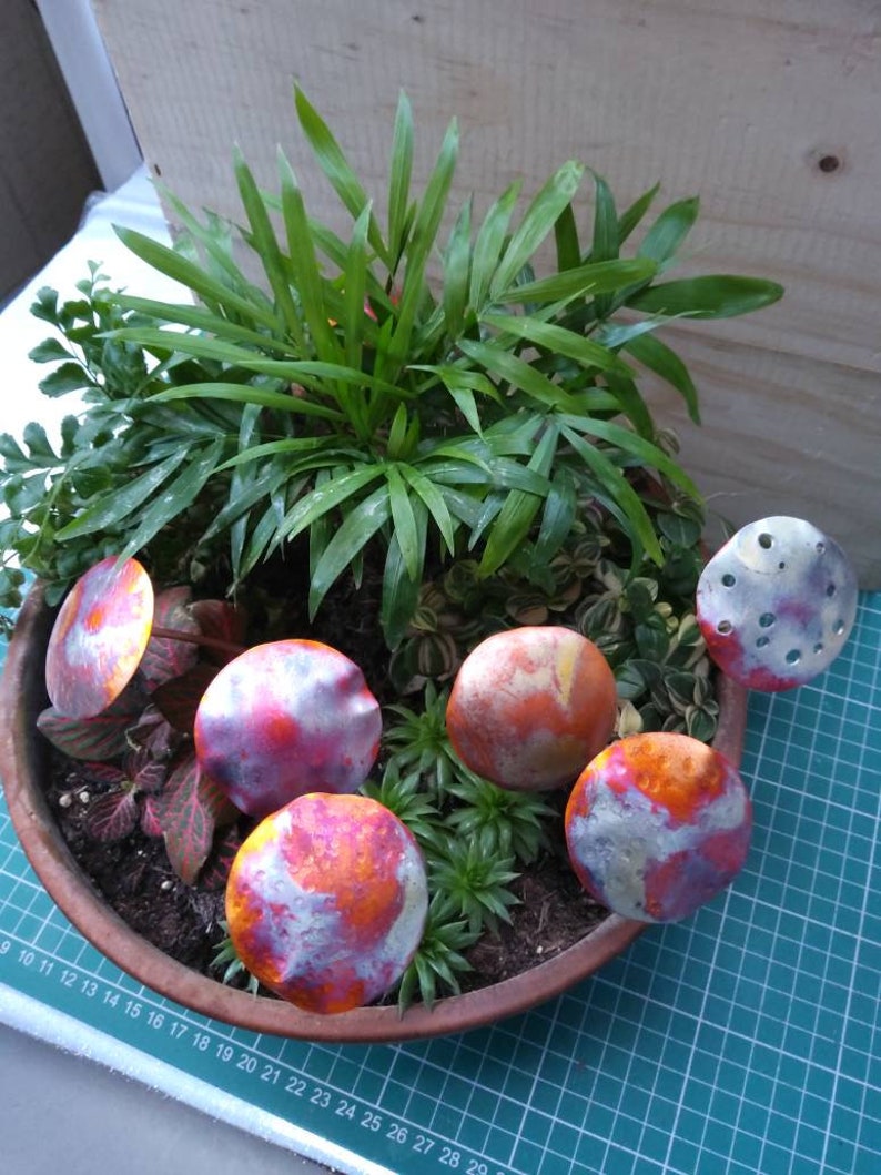 May include: Five small, metallic, mushroom-shaped ornaments in shades of orange, pink, and white, arranged in a terracotta pot with green plants.