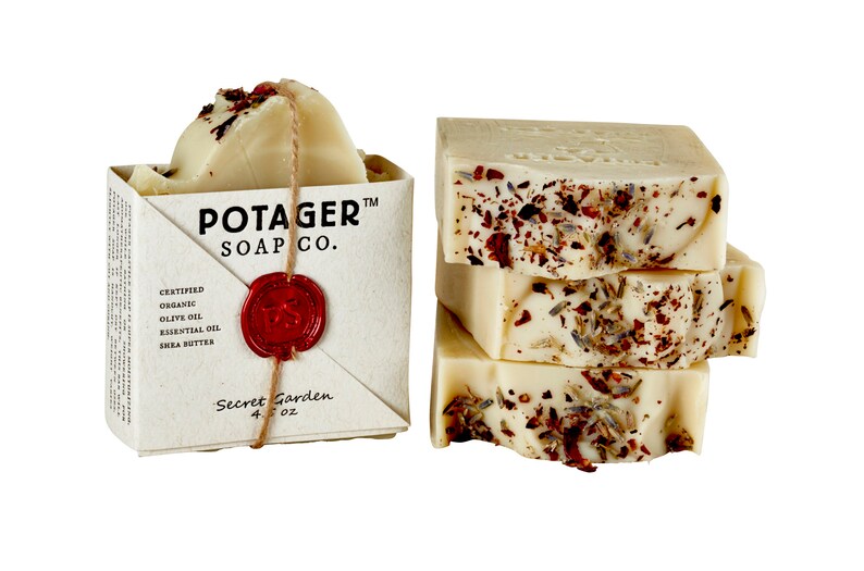 May include: A bar of white soap with dried flowers and herbs, wrapped in a brown paper envelope with the text "Potager Soap Co." and "Secret Garden 4 oz".