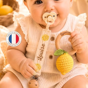 Può includere: Un bambino con un ciuccio e un anello per la dentizione in legno a forma di limone. La clip del ciuccio è ricamata con fette di limone e il nome "Louise". Il prodotto è contrassegnato come "Fabriqué en France".
