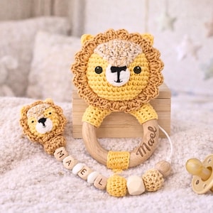 May include: A handmade crochet lion-themed baby set, including a teether, pacifier clip, and pacifier. The lion's mane is a light brown, with a yellow face and wooden accents. The pacifier is a light yellow color.