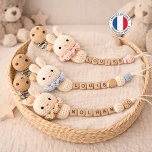 May include: Three handmade baby pacifier clips with crochet bunny heads, each with a different colored collar. The names "LOUISE", "LOUIS", and "NOLHAN" are spelled out with wooden letter beads. The pacifier clips are in a woven basket.