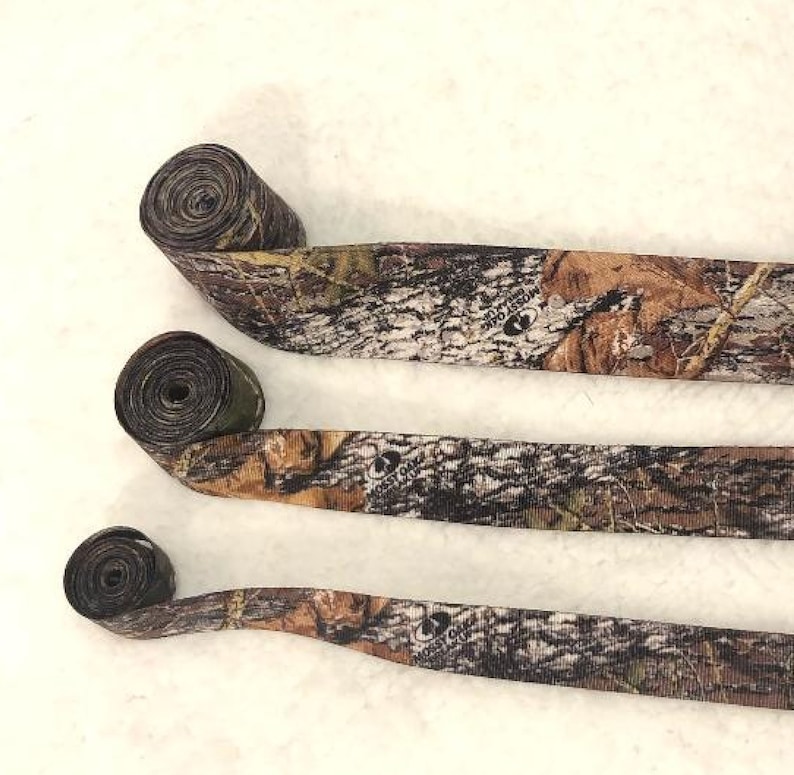 May include: Three rolls of camouflage patterned ribbon. The ribbon is brown, green and black with a white background. The ribbon has the words "Mossyoak" printed on it.