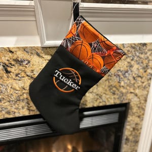 May include: Black Christmas stocking with a basketball theme. The top of the stocking features a pattern of orange basketballs and nets. The stocking has the name "Tucker" embroidered in orange.