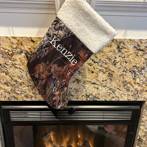 May include: A Christmas stocking with a camouflage pattern and a white sherpa cuff. The name "Kenzie" is printed in white script on the front. The stocking is hanging above a fireplace.