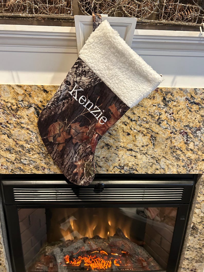 May include: A Christmas stocking with a camouflage pattern and a white sherpa cuff. The name "Kenzie" is printed in white script on the front. The stocking is hanging above a fireplace.