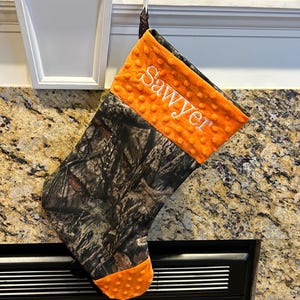 May include: A camouflage and orange Christmas stocking with the name "Sawyer" embroidered on the orange fabric.
