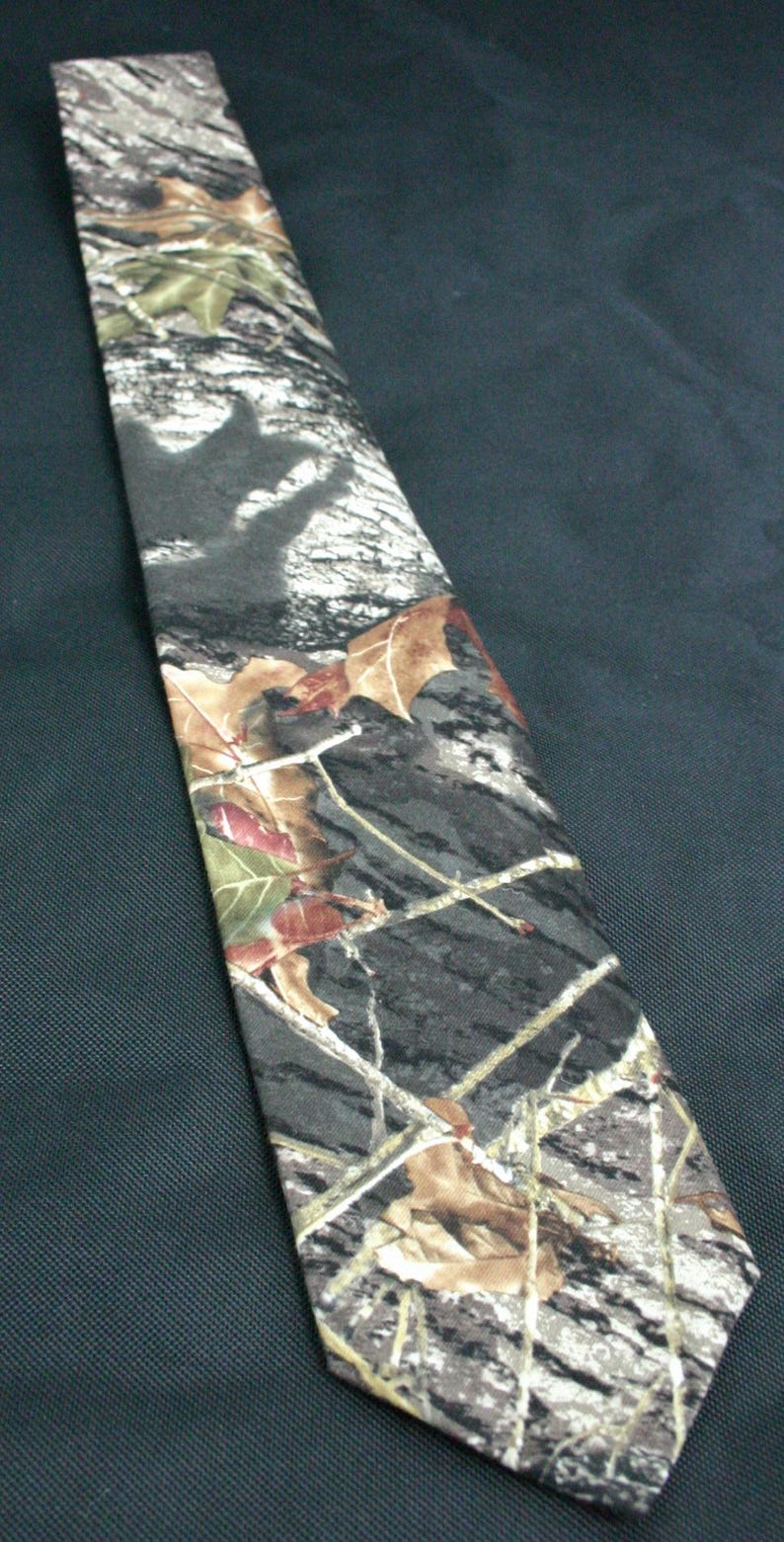 May include: A camouflage patterned tie with brown, green, and white colours. The tie has a leaf and branch design.