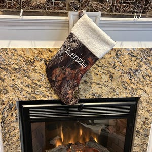 May include: A Christmas stocking with a camouflage pattern and a white, fluffy cuff. The name "Kenzie" is printed in white on the front. The stocking is hanging above a fireplace.