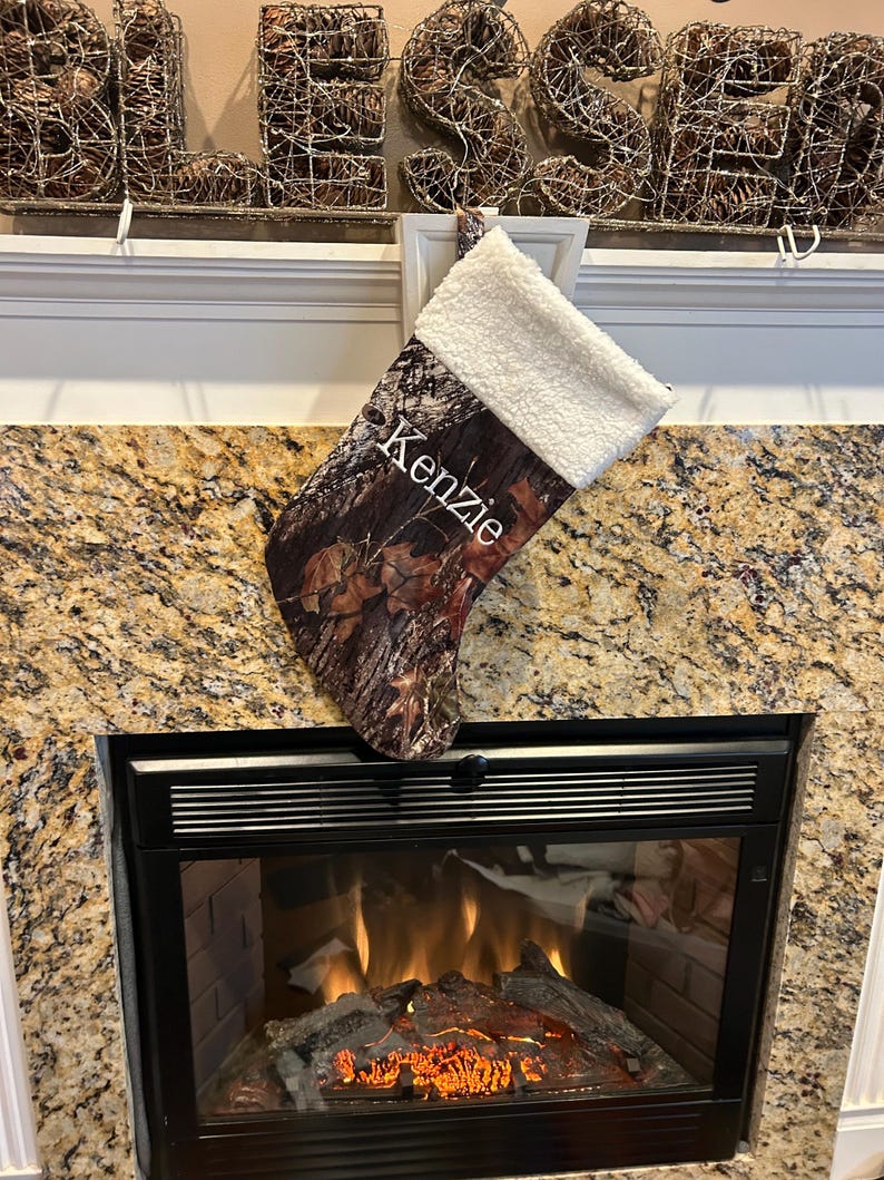 May include: A Christmas stocking with a camouflage pattern and a white, fluffy cuff. The name "Kenzie" is printed in white on the front. The stocking is hanging above a fireplace.