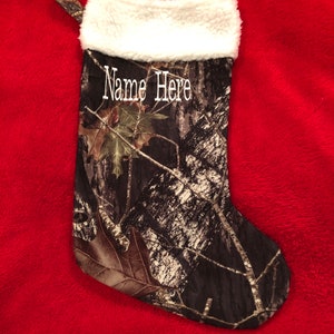 May include: Camouflage Christmas stocking with a white faux fur cuff and a loop for hanging. The stocking features the text "Name Here" in white script. The background is a red, plush fabric.