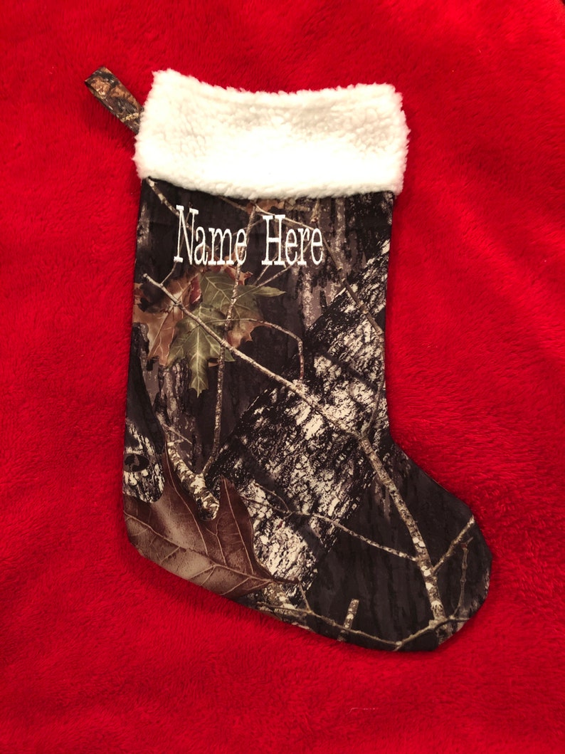May include: Camouflage Christmas stocking with a white faux fur cuff and a loop for hanging. The stocking features the text "Name Here" in white script. The background is a red, plush fabric.