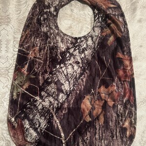 May include: A camouflage-patterned baby bib with a neck opening. The bib features a design of tree bark, branches, and leaves in shades of brown, green, and white. The bib is made of a soft, flexible material.