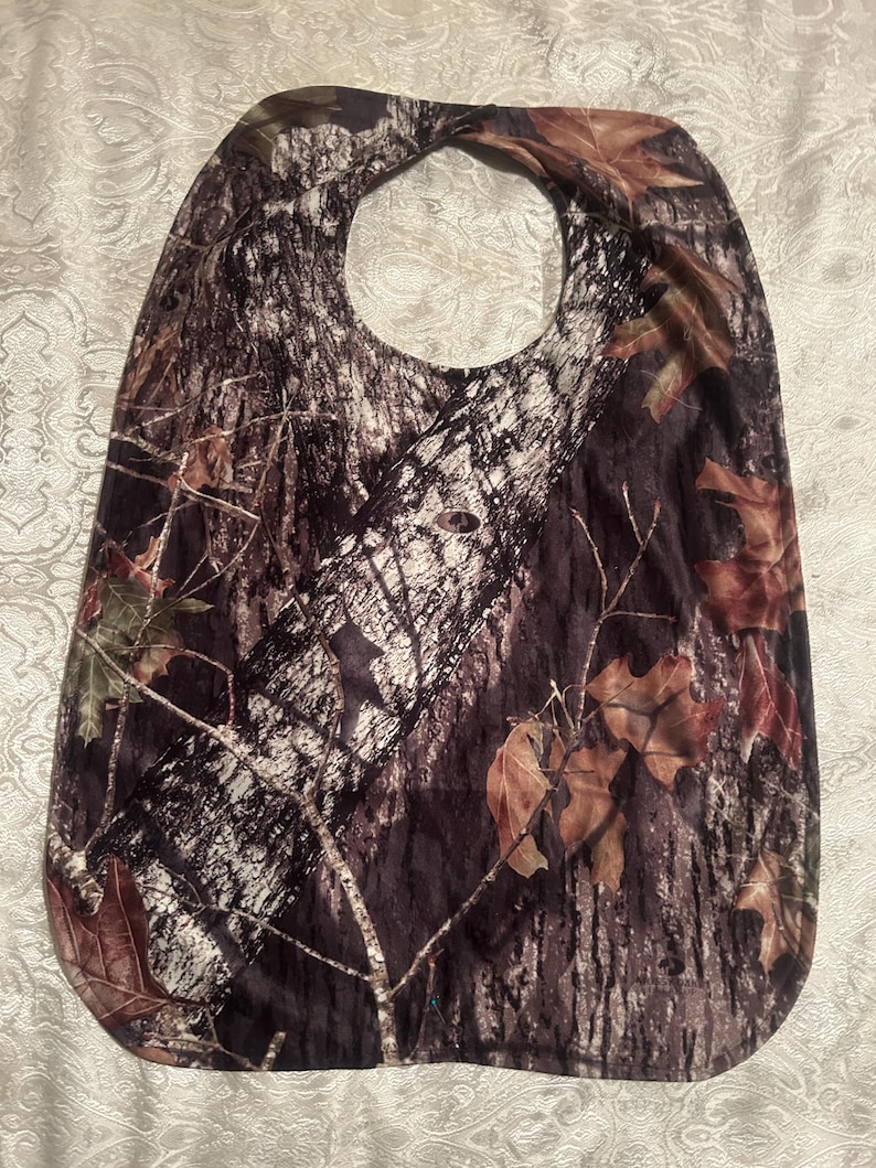 May include: A camouflage-patterned baby bib with a neck opening. The bib features a design of tree bark, branches, and leaves in shades of brown, green, and white. The bib is made of a soft, flexible material.