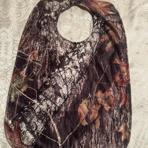 May include: A camouflage-patterned bib with a neck opening. The bib features a brown, green, and white tree bark design with scattered brown leaves. The bib is made of a soft, flexible material.