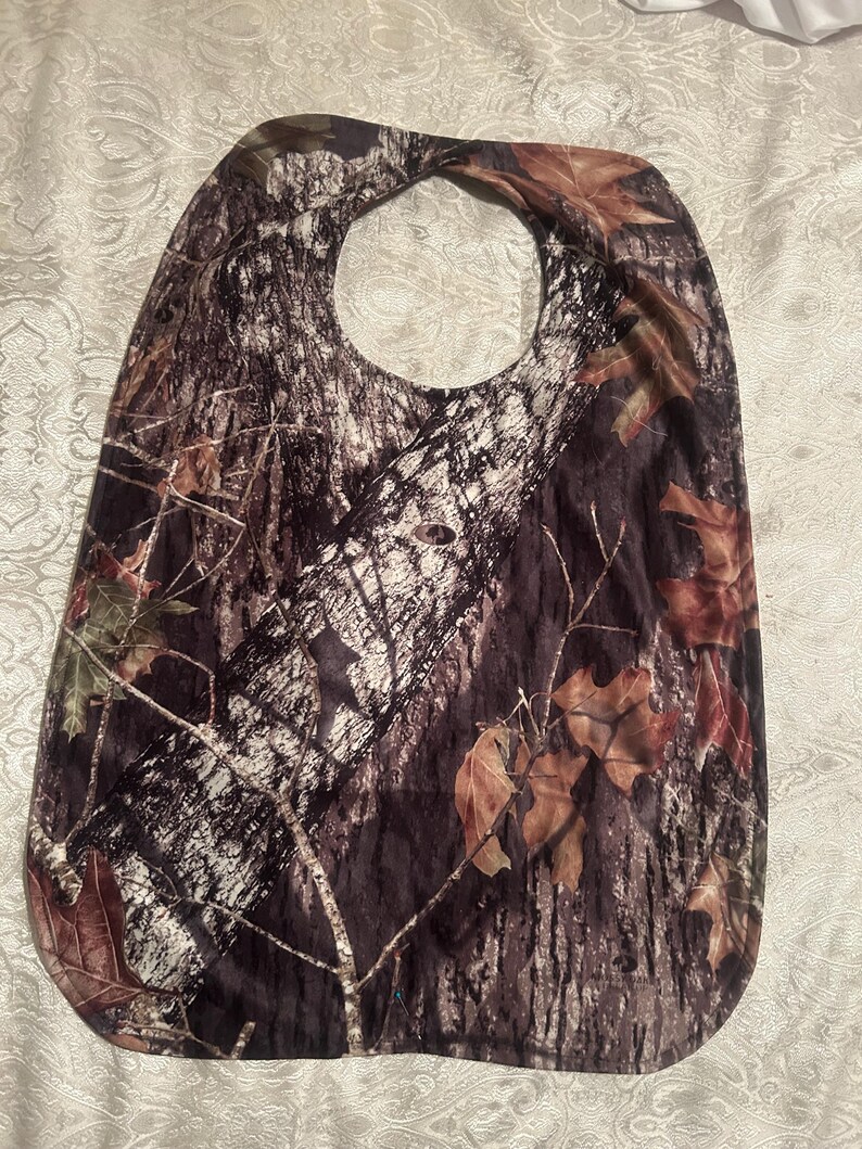 May include: A camouflage-patterned bib with a neck opening. The bib features a brown, green, and white tree bark design with scattered brown leaves. The bib is made of a soft, flexible material.