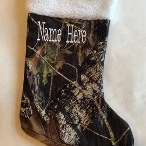 May include: A Christmas stocking with a camouflage pattern, featuring a white sherpa cuff. The words "Name Here" are embroidered in white. A matching fabric loop is attached for hanging.