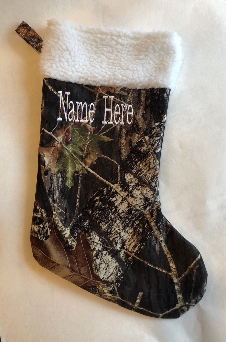 May include: A Christmas stocking with a camouflage pattern, featuring a white sherpa cuff. The words "Name Here" are embroidered in white. A matching fabric loop is attached for hanging.
