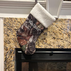 May include: A Christmas stocking with a camouflage pattern, featuring the name "Walker" in white script. The top of the stocking is trimmed with fluffy white material, and it hangs from a white hook. The stocking is displayed near a fireplace.