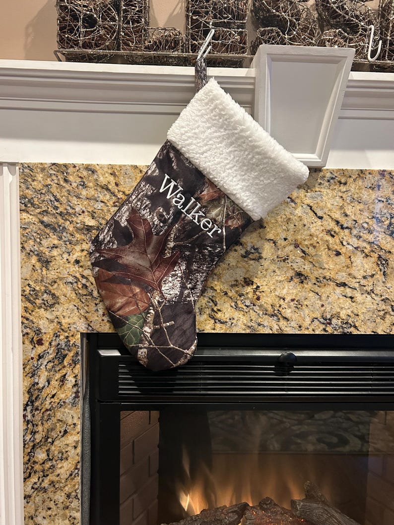 May include: A Christmas stocking with a camouflage pattern, featuring the name "Walker" in white script. The top of the stocking is trimmed with fluffy white material, and it hangs from a white hook. The stocking is displayed near a fireplace.