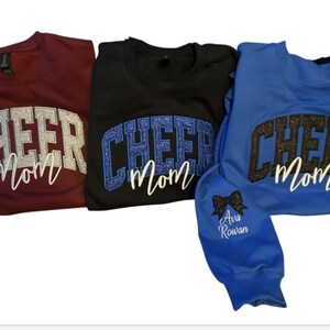 Puede incluir: Tres sudaderas con el texto "Cheer Mom" en letras de purpurina. Las sudaderas son granate, negra y azul.