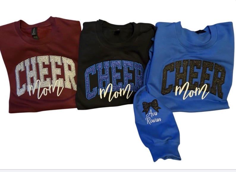 Puede incluir: Tres sudaderas con el texto "Cheer Mom" en letras de purpurina. Las sudaderas son granate, negra y azul.