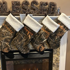 May include: Four camouflage Christmas stockings with white faux fur trim hang above a fireplace. Each stocking has a name embroidered in orange: Daddy, Mommy, Brandon, and Easton. The word "Blessed" is displayed above the stockings.