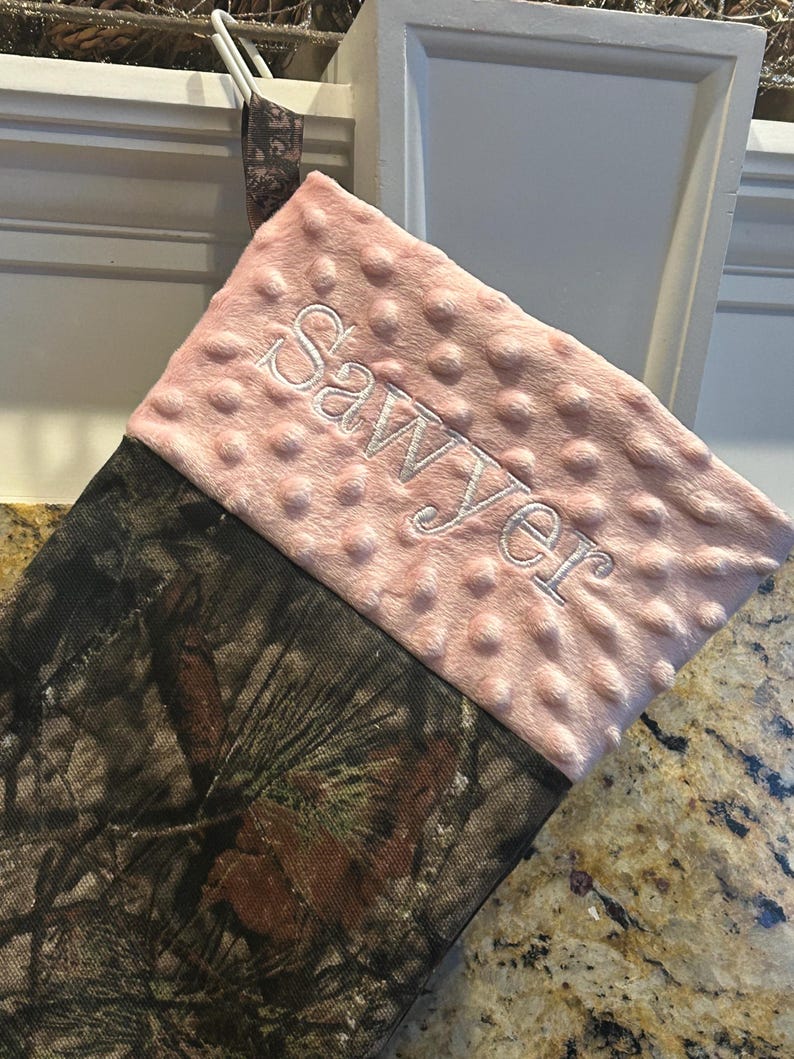 May include: A pink and brown camouflage Christmas stocking with the name "Sawyer" embroidered on the pink cuff.