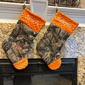 May include: Two personalized Christmas stockings with a camouflage pattern and orange trim. The stockings are hung on a fireplace mantel and are embroidered with the names "Jacob" and "Sawyer".